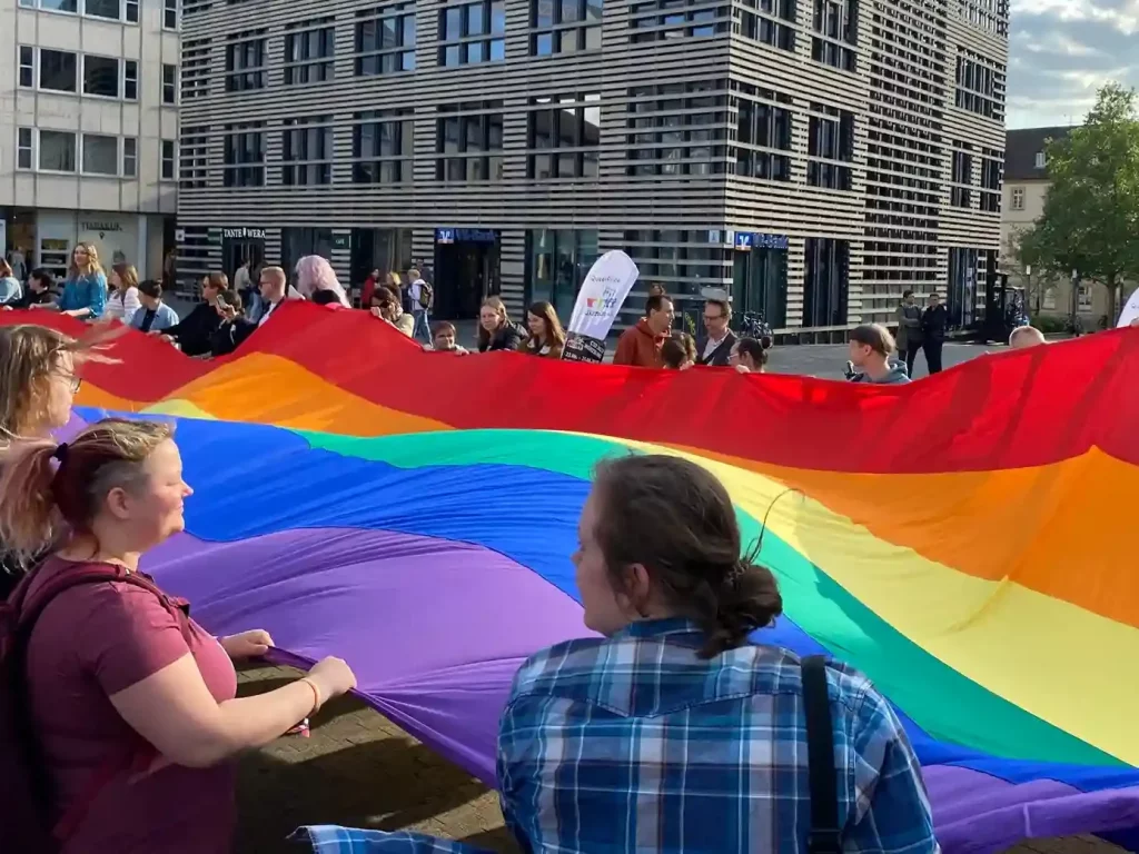 Menschen halten eine große Regenbogenflagge auf dem Marktplatz in Würzburg während einer queeren Aktion des WuF-Zentrums.